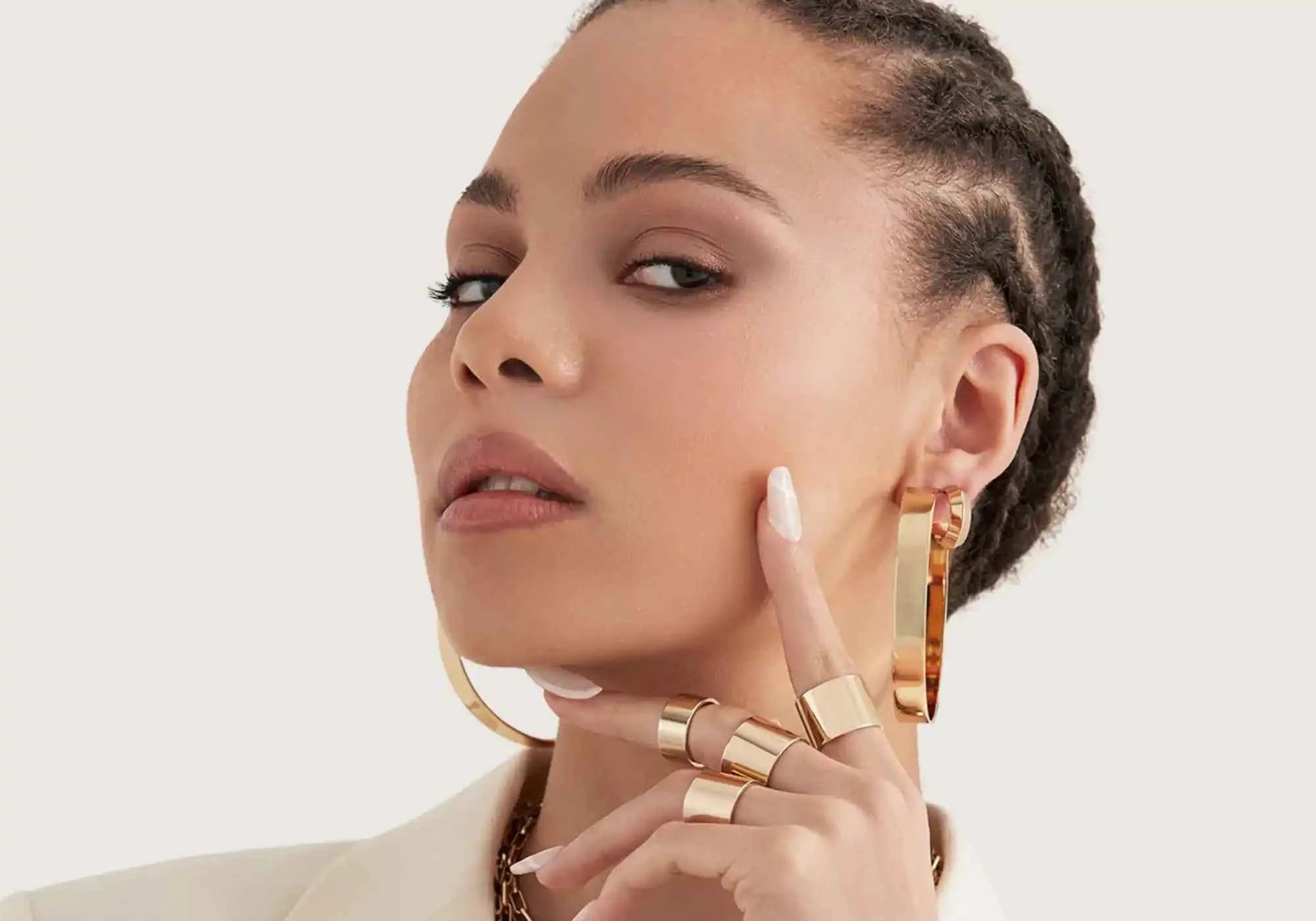 Woman wearing gold hoop earrings and rings on a plain background